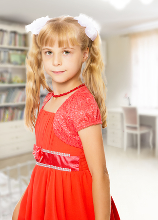 A very beautiful little girl with long, blonde ponytails on her head in a bright orange dress.In the background children's room where there are shelves with books and toys.の写真素材