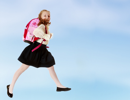 Pretty little girl schoolgirl in white blouse and black skirt to hurry to school. She jumps over the obstacle.On the pale blue background.の写真素材