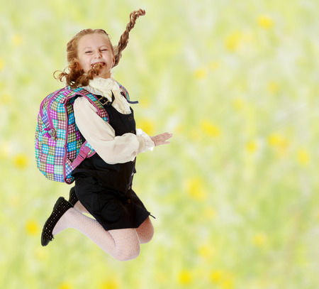 Happy girl schoolgirl in black dress and white shirt and a knapsack on his shoulders. She jumps, hands up. Turning sideways to the camera.Summer white green blurred background.の写真素材