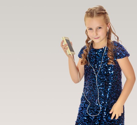 Elegant little girl in a festive, glittering, blue dress, holding a mobile phone . She listens to music through headphones.On a gray background.の写真素材