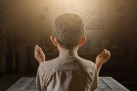 Young muslim boy in prayer cap and arabic clothes with rosary beads and holy Koran book praying to...