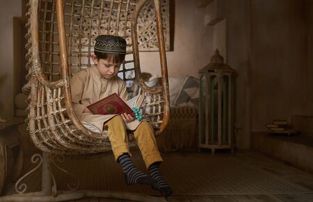 Small muslim boy in prayer cap and arabic clothes with rosary beads reading holy Koran book praying...