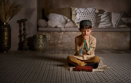 Boy in prayer cap and arabic clothes with rosary beads and holy Koran book praying to Allah, ramadan kareem concept young kid spiritual peaceful moment inside eastern traditional interiorの写真素材