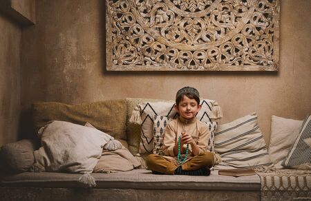 Little muslim boy in prayer cap and arabic clothes with rosary beads reading holy koran book praying to Allah, prophet Muhammad holy spirit religion symbol concept inside eastern traditional interiorの写真素材