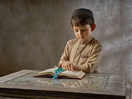 muslim boy in prayer cap and arabic clothes with rosary beads reading holy koran book praying to...
