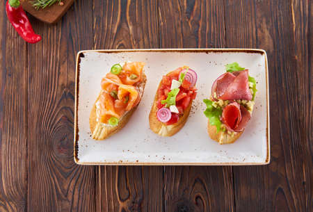 Bruschetta with various toppings, variety of small sandwiches with salmon red fish, fresh vegetables, tomatos and herbs on wooden table, diet food ingredients background.の写真素材