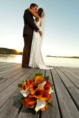 wedding bouquet in foreground and newlyweds kissing behind at sunsetの写真素材