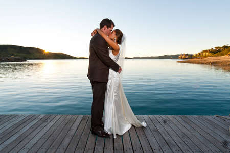 Newlyweds kissing on a jetty at sunset with beautiful water behindの写真素材