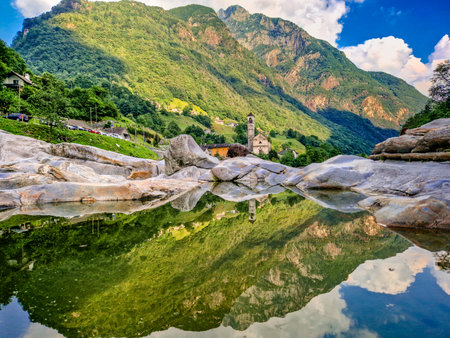 A picturesque photo captures a serene mountain village and a church reflected in a calm river, surrounded by lush green hills and towering mountains under a vibrant blue skyの写真素材