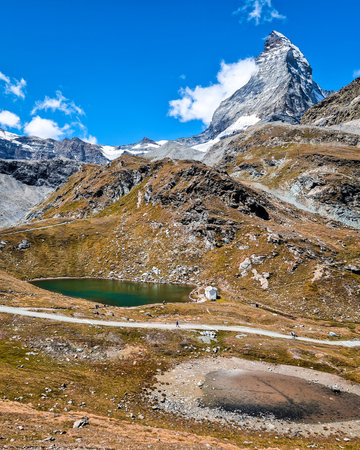 A scenic mountain landscape featuring a small alpine lake and rugged terrain, with the iconic Matterhorn peak towering under a bright blue sky, dotted with a few cloudsの写真素材