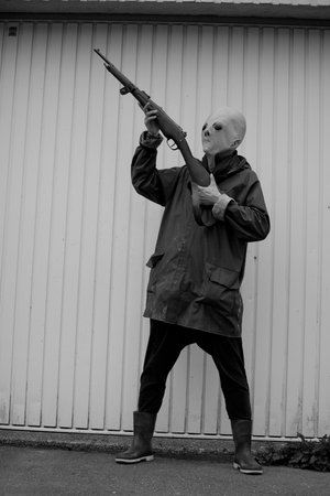 black and white photography. Woman wearing an alien mask hunting with shotgun in front of a garage...