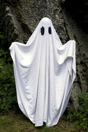 A spooky white ghost covered by a sheet with slits over the eyes standing in front of a rock.の写真素材