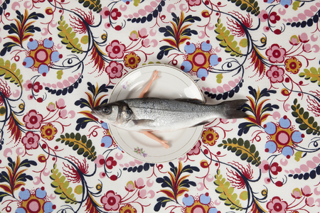 A bass fish with arms doll inside a flower plate hidden on a flowery fabric. Camouflage game and anthropomorphism. Minimal color still life photography.の写真素材