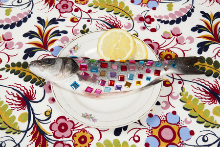 A bass fish covered with colorful precious stones inside a flower plate hidden on a pop floral patterned fabric background. Minimal quirky color still life photography.の写真素材