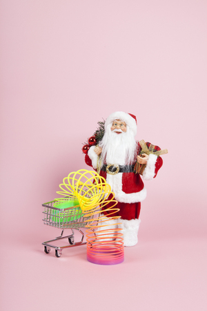a santa claus figurine plush placed in front of a shopping trolley with rainbow spring inside on a pink background. Color harmony. Minimal still life color photographyの写真素材