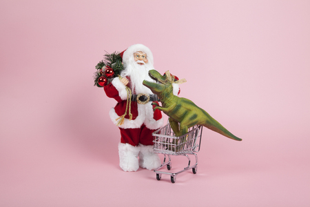 a santa claus figurine plush placed in front of a shopping trolley with tyrannosaurus plastic toy inside on a pink background. Color harmony. Minimal still life color photographyの写真素材