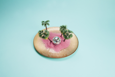 a golden plate with pink sand and mini plastic palm trees inside as well as a disco ball on a turquoise background. Minimal still life color photographyの写真素材
