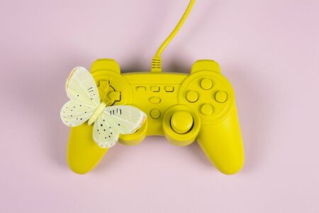 a yellow painted video game controller and a yellow butterfly on it on a plain pink background....