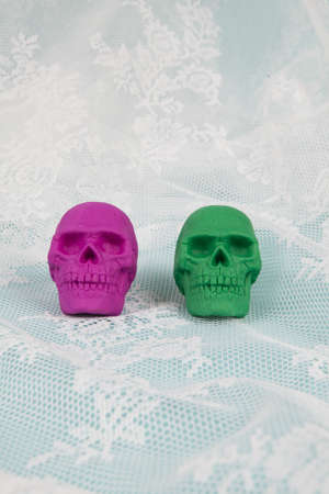 Two plastic skulls, one pink and one green, placed on a lace curtain that looks like sea foam. Minimal and creative color still life photographyの写真素材