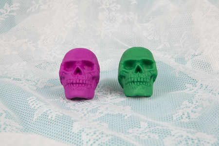 Two plastic skulls, one pink and one green, placed on a lace curtain that looks like sea foam. Minimal and creative color still life photographyの写真素材
