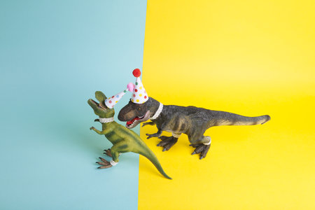 a couple of dinosaurs, dressed up in lace and a paper party hat, at a birthday party against a yellow and blue background. Minimal creative still life colorful photographyの写真素材