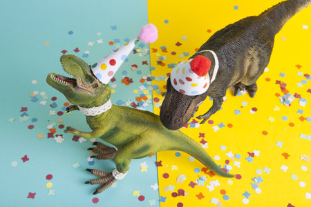 a pair of dinosaurs celebrating on a confetti carpet, dressed in lace and a paper party hat, at a birthday party against a yellow and blue background. Minimal creative still life colorful photographyの写真素材