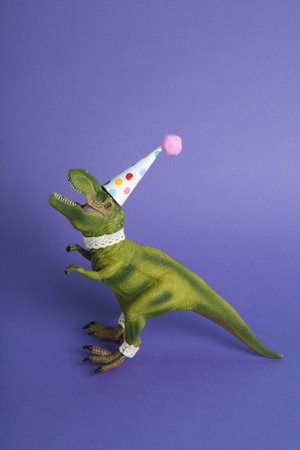 a dinosaur, dressed up in lace and a paper party hat, at a birthday party against a purple background. Minimal creative still life colorful photographyの写真素材