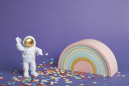 an astronaut figurine in a spacesuit exploring for treasure a plain purple background on a confetti carpet and in front of a plastic rainbow. Minimal creative still life colorful photographyの写真素材