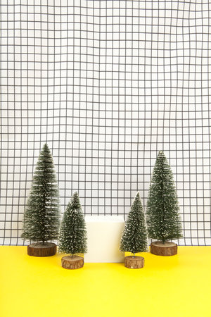 a white wooden cube surrounded by several miniature christmas trees on a black and white grid background. empty object display stand. Minimal still life photographyの写真素材