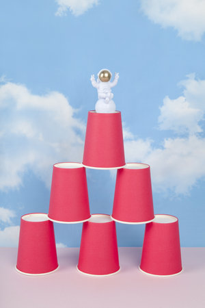 a white astronaut figurine with gold helmet on a pyramid of red paper cups, set against a background of blue summer sky with white clouds.Vivid color and minimal pop art photographyの写真素材