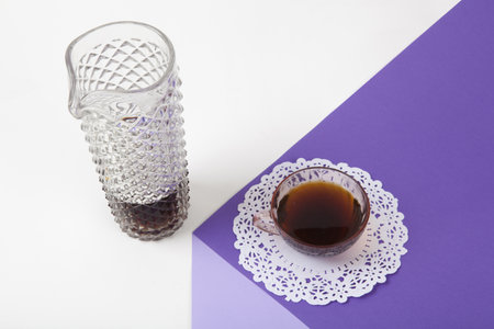 a violet cube made with colored paper to create a 3D optical illusion. On top is a vintage pink coffee cup with a white lace doily and a glass jug of coffee imitation, fake crystal. Vivid colors and minimal pop art photography.の写真素材