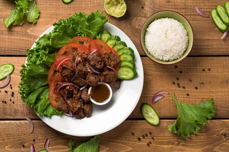 Bo Luc Lac dish on wooden background. Also named Lok Lak in cambodia or Shaking Beef in America. Tasty marinated sauteed beef cubes are served with fresh lettuce, tomatoes, cucumbers, onions and rice.の写真素材