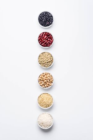Various legumes and grains top view. Bowls of staple food ingredients like chickpeas, red kidney beans, green lentils, black turtle beans, jasmine rice and basmati rice on white background from above.の写真素材