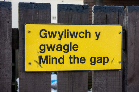 Metal mind the gap warning sign attached to a fence at rear of railway platform