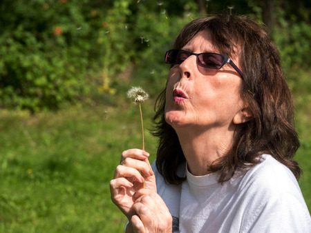Attractive mature lady blowing Dandelion seeds to tell the timeの写真素材