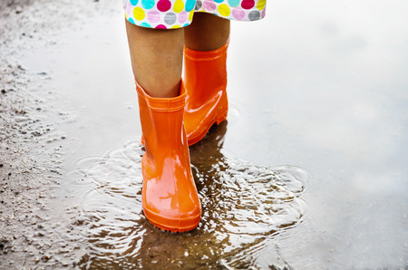 Child wearing orange rain boots walking into a puddle. Close up