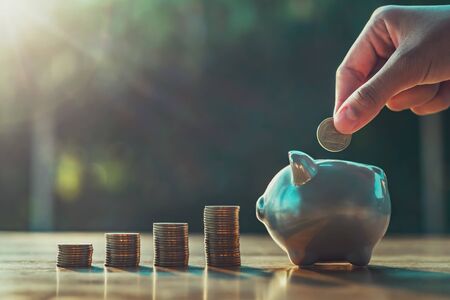 hand puting coins in piggy bank with money stack on desk
