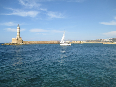 Sailing White Yacht near the Lighthouse at the Old Venetian Harbour of Chania, Crete Island, Greeceの写真素材