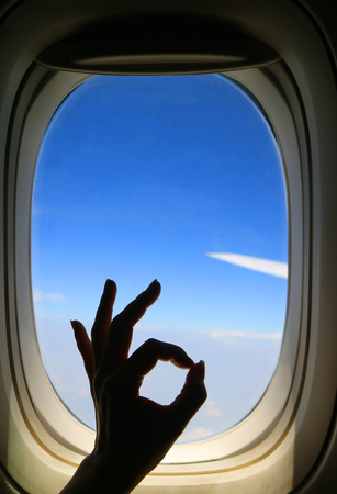 Silhouette of female's hand posing OK sign against airplane window with vibrant blue sky and airplane wing during her happy flightの写真素材