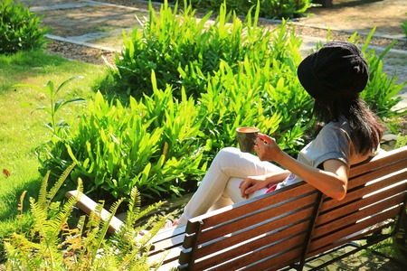Young woman relaxing on wooden bench in vibrant green garden holding a cup of hot coffee in the morningの写真素材