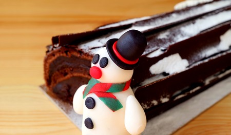 Close-up of a Cute Snowman Marzipan with Blurred Chocolate Yule Log Cake in Background Isolated on Wooden Tableの写真素材