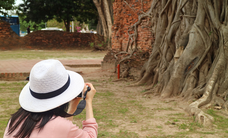 Female Tourist Taking Pictures of Buddha Image's Head Trapped in Bodhi Tree Roots, Wat Mahathat Ancient Temple, Ayutthaya Historical Park, Archaeological site in Thailandの写真素材