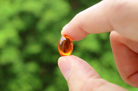 Closeup Man's Fingers Holding a Transparent Supplement Gelcap Against Blurry Green Foliageの写真素材