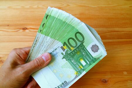 Bunch of 100 EURO Banknotes in Man's Hand with Wooden Table in the Backdropの写真素材