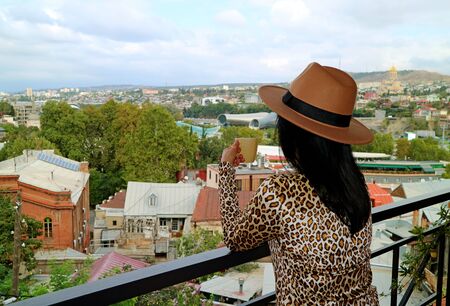 Woman in hat holding a cup of coffee while enjoy the aerial view of Tbilisi, the capital of Georgiaの写真素材