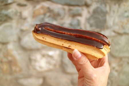 Hand Holding a Chocolate Eclair with Blurry Stone Wall in Background