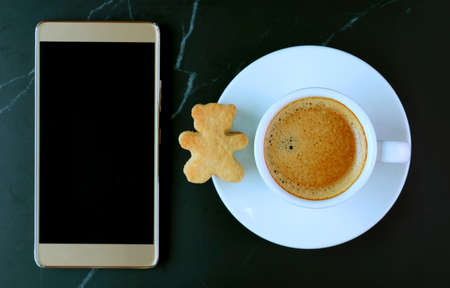 Empty Screen Mobile Phone with a Cup of Coffee and Teddy Bear Shaped Cookie on Black Marble Table