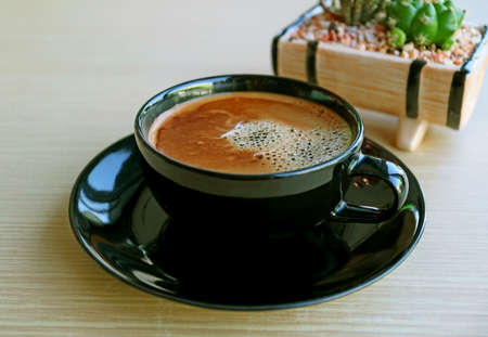Closeup a cup of frothy hot coffee with blurry potted plant on wooden table