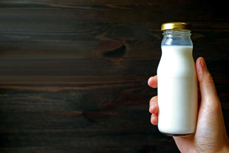 Man's hand holding a glass bottle of milk against dark brown wooden backgroundの写真素材