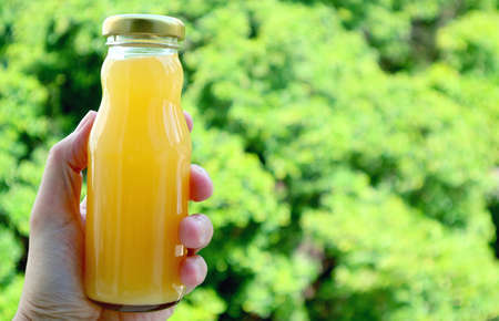 Glass bottle of orange juice in man's hand with blurry green foliage in the backdrop